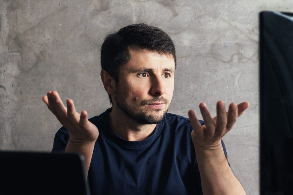 Man with dark hair and stubble raises both hands in front of a laptop, looking puzzled against a gray concrete wall.