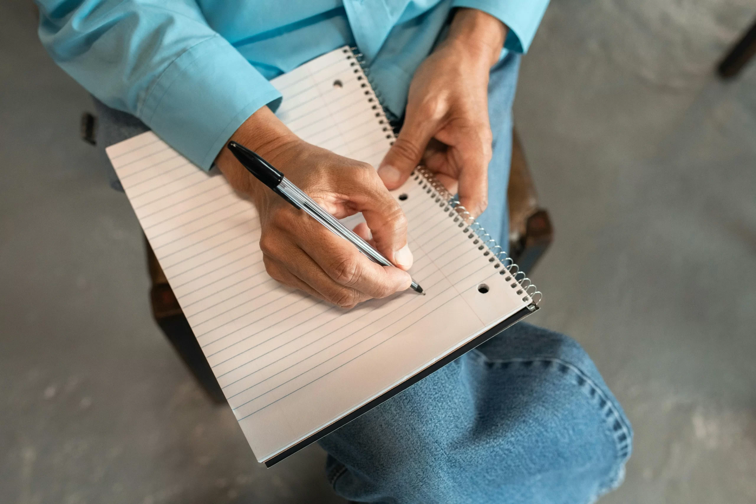 Person wearing a blue shirt writes in a spiral notebook resting on their knee.